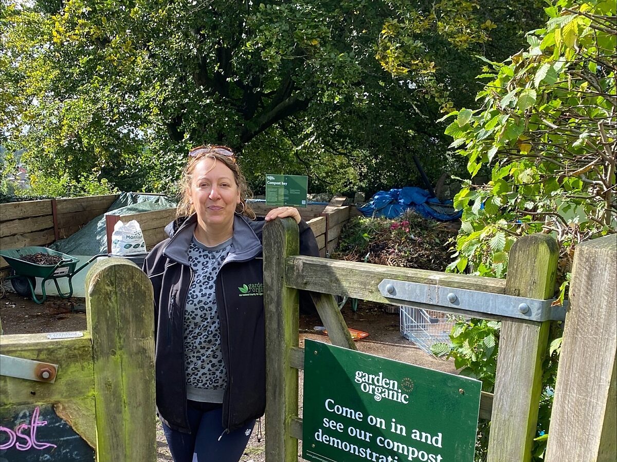 Project Coordinator Frankie Kennet, stands at the gate of a compost demonstration site, next to a sign that reads "come on in and see our compost demonstration site"